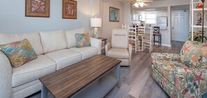 Ein Wohnzimmer mit einer Couch, einem Couchtisch, einem Sessel und einem gemusterten Ohrensessel. Im Hintergrund ist ein Essbereich mit Tisch und Stühlen zu sehen.