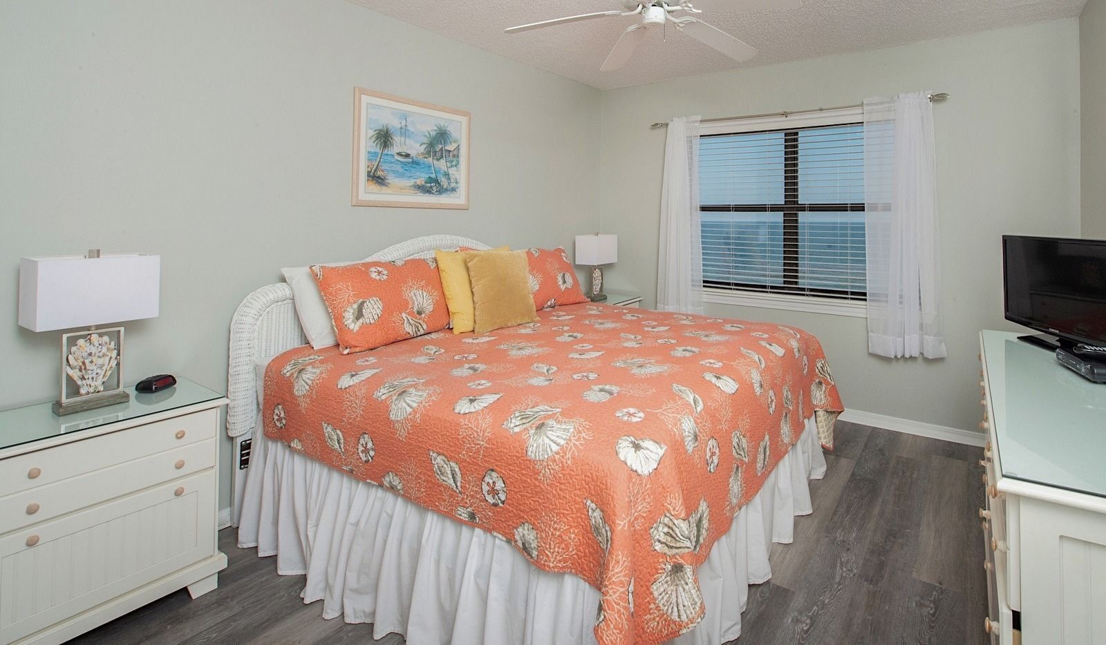 A bedroom with a bed, a nightstand, a TV, a window with blinds, and a ceiling fan.