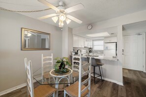 Een eetruimte met een glazen tafel, witte stoelen, een plafondventilator en een keuken op de achtergrond.