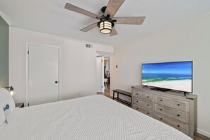 Ein Schlafzimmer mit einem Bett, einem Deckenventilator, einem Fernseher auf einer Holzkommode und Blick auf den Strand auf dem Bildschirm.