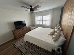 Ein Schlafzimmer mit einem Bett, einem Fernseher auf einer Holzkommode, einem Deckenventilator und einem Fenster mit Jalousien.