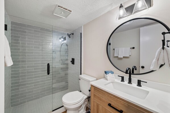 A modern bathroom with a glass-enclosed shower, a white toilet, a wooden vanity, and a round mirror. - Sunswept 707 by Liquid Life (Orange Beach)