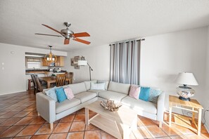 A living room with a sectional sofa, a coffee table, a dining area with a kitchen in the background, and a ceiling fan. - Sunswept 801 Penthouse by Liquid Life (Orange Beach)