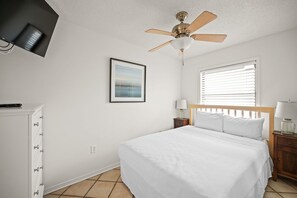 A hotel room with a bed, a nightstand, a ceiling fan, a window with blinds, a framed picture on the wall, and a television mounted on the wall. - Sunswept 801 Penthouse by Liquid Life (Orange Beach)