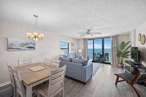A modern living room with a dining area, a ceiling fan, and a view of the ocean through large windows. - Sunswept 801A by Liquid Life (Orange Beach)