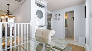 A modern interior with a glass table, a white chair, a washing machine, a clock, and a door leading to another room. - Sunswept 804 by Liquid Life (Orange Beach)