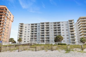 Un immeuble résidentiel à plusieurs étages doté de balcons, une plage de sable au premier plan et un ciel dégagé.