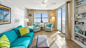 Ein Wohnzimmer mit einem salbeingrünem Sofa, einem Couchtisch, einem Deckenventilator, einem Bücherregal und Blick ins Freie durch ein großes Fenster.