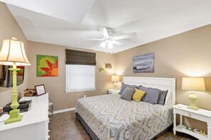 A bedroom with a bed, two bedside tables, a ceiling fan, a window with blinds, and wall art. - Sunswept 806 Penthouse by Liquid Life (Orange Beach)