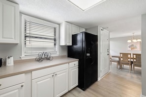 A kitchen with white cabinets, a black refrigerator, a window with blinds, and a dining area with wooden chairs and a table. - Sunswept 807 Penthouse by Liquid Life (Orange Beach)