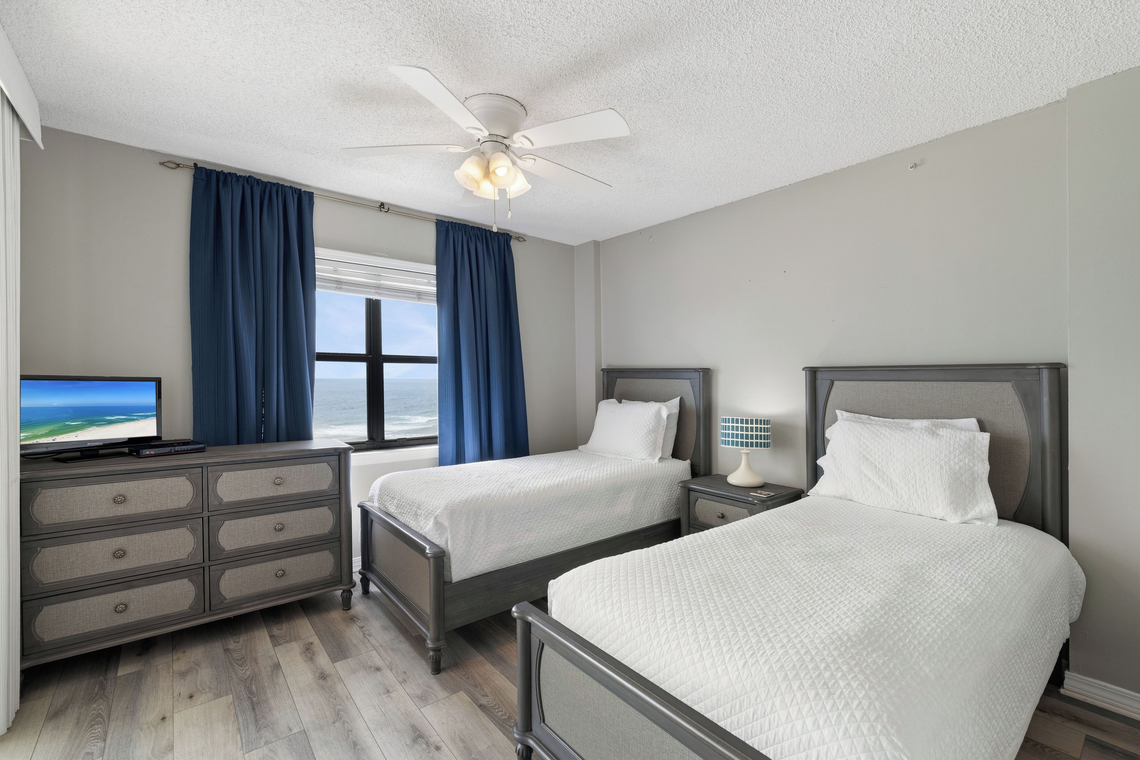 Quarto de hotel com duas camas, televisão, penteadeira e vista para o oceano.