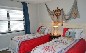 A room with two beds, a wall-mounted ship's wheel decoration, and a lamp on a bedside table.