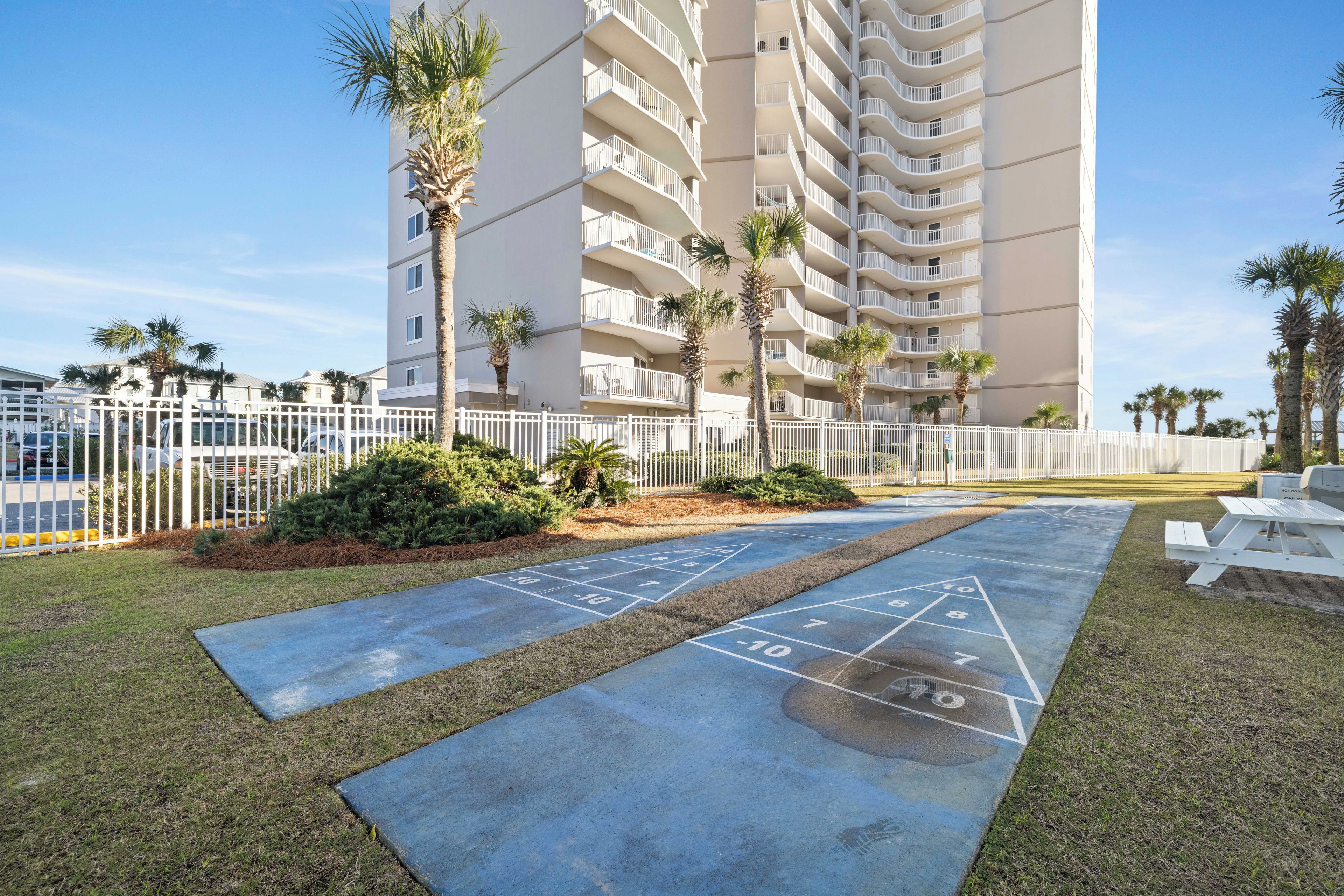 A courtyard with a shuffleboard court, palm trees, and a white fence.