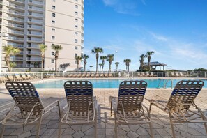 A poolside area with lounge chairs, a pool, palm trees, and a high-rise building in the background. - Seaside Beach & Racquet 4612 by Liquid Life (Orange Beach)