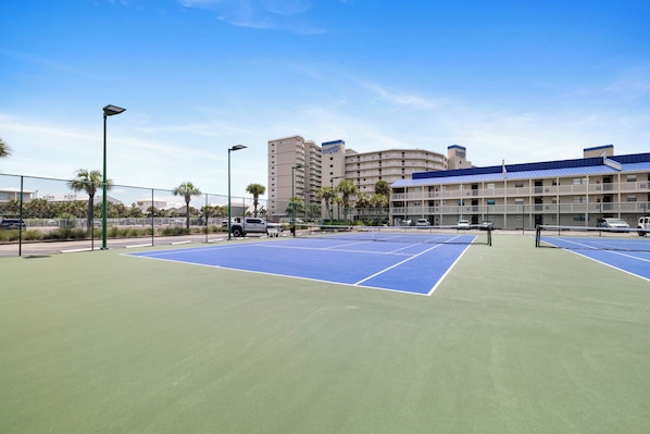 Ein Tennisplatz mit Netz, umgeben von einem Zaun, und ein Hotelgebäude im Hintergrund.