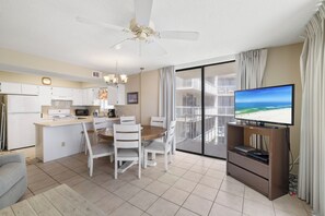 A modern kitchen with white cabinets, a microwave, and a coffee maker. There is a dining area with a wooden table and white chairs. A television is on a stand to the right. - Seaside Beach & Racquet 4612 by Liquid Life (Orange Beach)