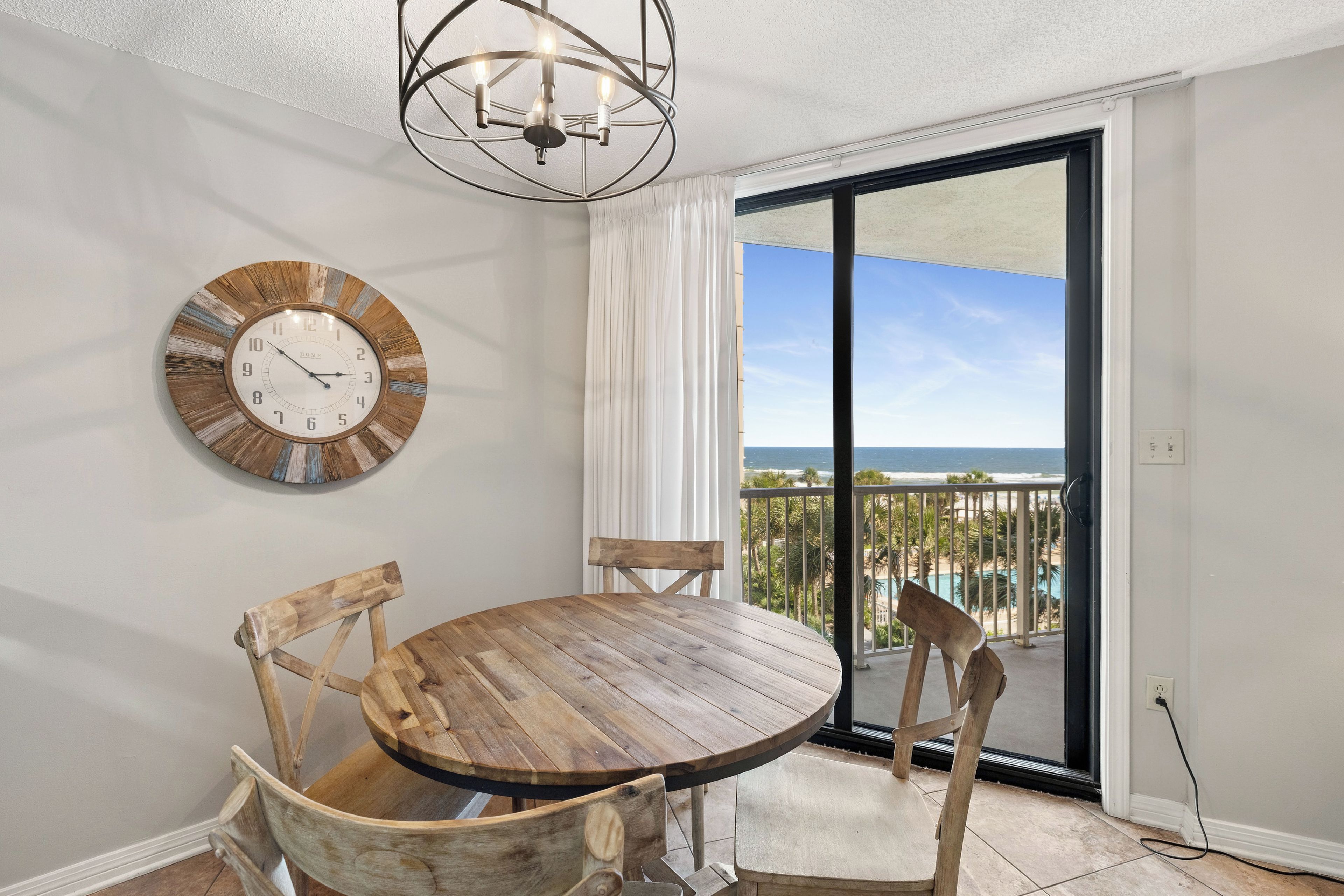 A dining area with a wooden table and chairs, a wall clock, and a view of the ocean through a sliding glass door.