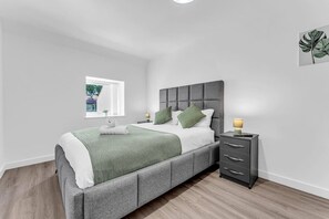 A modern bedroom with a grey upholstered bed, white bedding, green pillows, a bedside table with a lamp, and a window with a view of greenery. - Spacious & Modern Anfield Flat | WiFi | Sleeps 4 (Anfield)