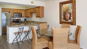 A kitchen with wooden cabinets, a stainless steel refrigerator, a microwave, and a coffee maker. There is a glass island with a sink and a dining table with wicker chairs. A framed painting hangs on the wall. - Seaside Beach & Racquet 5915 by Liquid Life (Orange Beach)
