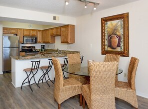 A kitchen with wooden cabinets, a stainless steel refrigerator, a microwave, and a coffee maker. There is a glass island with a sink and a dining table with wicker chairs. A framed painting hangs on the wall. - Seaside Beach & Racquet 5915 by Liquid Life (Orange Beach)