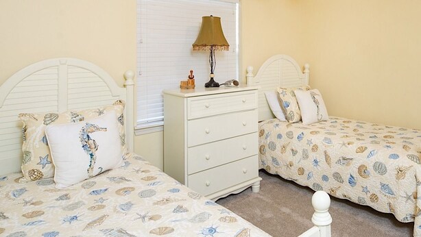 A bedroom with two beds, a nightstand, a lamp, and a window with blinds. - Seaside Beach & Racquet 5915 by Liquid Life (Orange Beach)