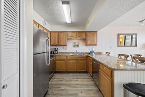 A kitchen with wooden cabinets, a stainless steel refrigerator, a granite countertop, and a window with a view. - Seaside Beach & Racquet 5915 by Liquid Life (Orange Beach)
