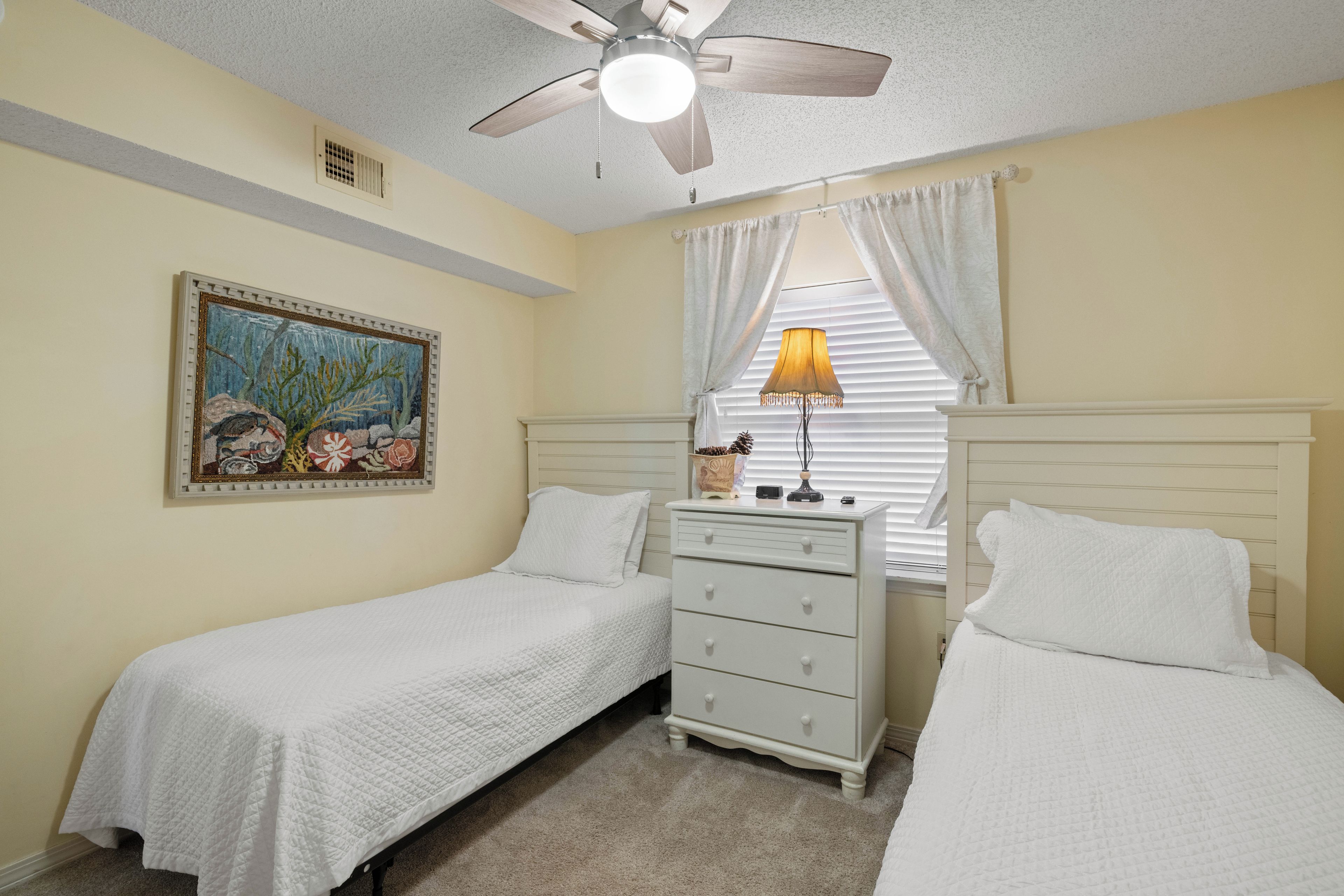 A bedroom with two single beds, a ceiling fan, a nightstand with a lamp, and a painting on the wall.
