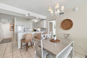 A modern kitchen with white cabinets, a dining area with a wooden table and wicker chairs, and a ceiling with a chandelier. - Shoalwater 604 by Liquid Life (Orange Beach)