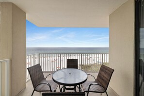 Varanda com mesa de vidro e cadeiras de vime, com vista para uma praia com guarda-sóis e pessoas.