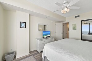 Une chambre d’hôtel avec un lit, un téléviseur posé sur une commode à plateau en verre, un ventilateur de plafond et une vue sur l’océan par la fenêtre.