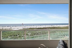 Blick auf einen Strand durch ein Fenster mit Geländer, eine Flagge und Strandstühle.