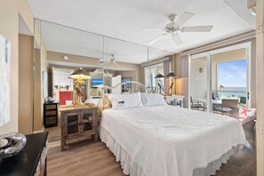 Une chambre d’hôtel avec un grand lit, un ventilateur de plafond, un bureau avec une lampe et une vue sur l’océan par la fenêtre.