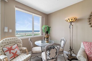 Une pièce avec vue sur la plage, une table ronde en verre, des chaises en osier, un lampadaire et un canapé recouvert d’un tissu à motifs léopard.