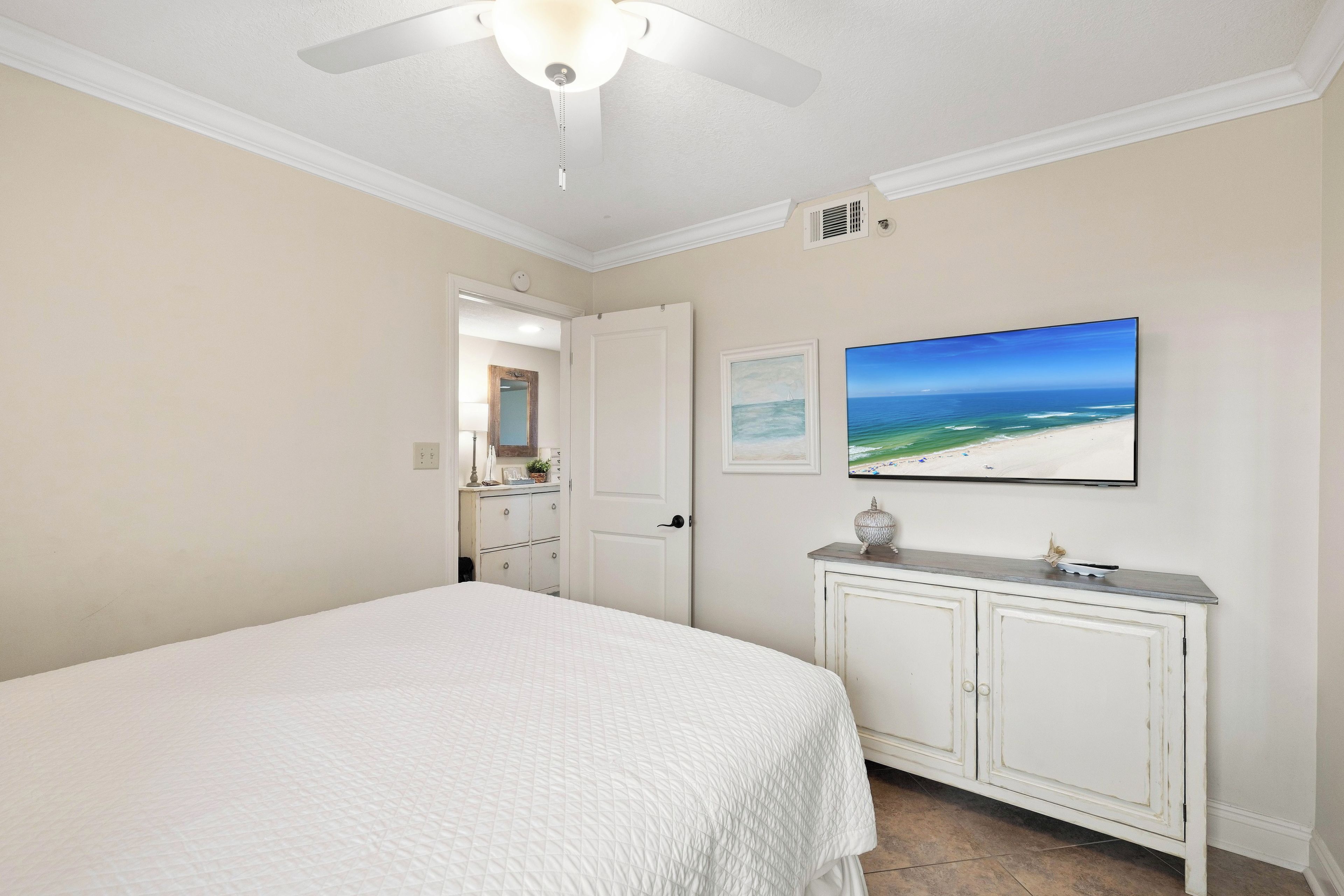Une chambre à coucher avec un lit, une commode, un ventilateur de plafond et un tableau représentant une scène de plage.