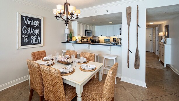 Uma sala de jantar com uma mesa branca posta para seis pessoas, rodeada por cadeiras de vime. Um candelabro pendurado sobre a mesa, e um letreiro na parede diz 'Vintage Cottage down by the SEA'.
