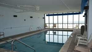 Piscina interna com água azul cristalina, prancha de salto e vista para a praia através de grandes janelas.