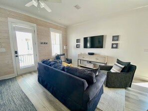 A living room with a dark blue sofa, a wooden floor, a flat-screen TV, and a window with blinds. - Kingfisher Cottage at Tannin by Liquid Life (Orange Beach)