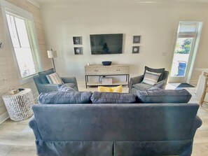 A living room with a grey sofa set, a flat-screen TV mounted on the wall, and a small cabinet below. - Kingfisher Cottage at Tannin by Liquid Life (Orange Beach)