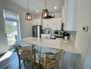 A modern kitchen with white cabinetry, a stainless steel refrigerator, a built-in oven, and a large island with two wooden chairs. - Kingfisher Cottage at Tannin by Liquid Life (Orange Beach)
