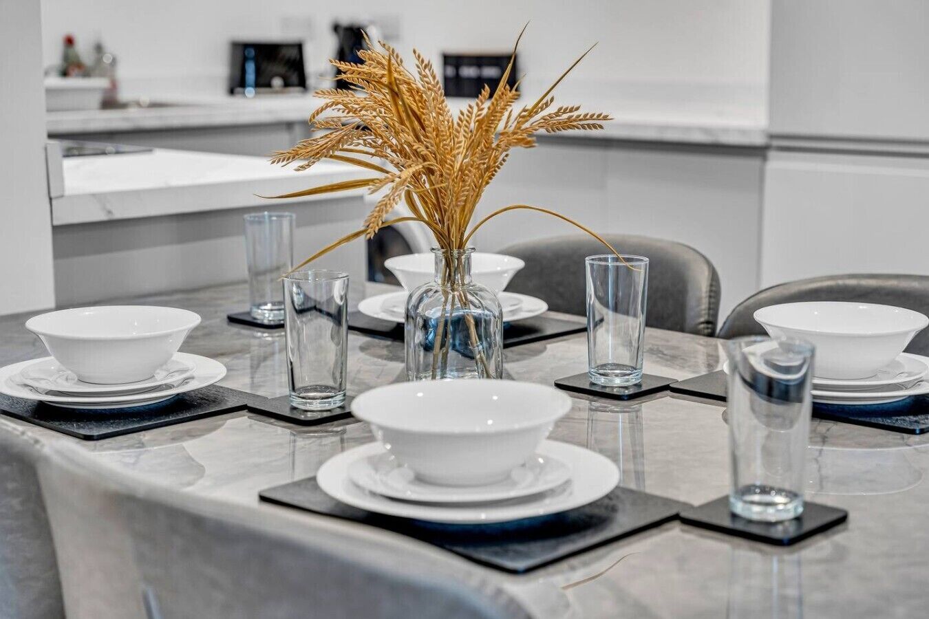 A modern dining table set with white plates, clear glasses, and a vase with dried wheat stalks.