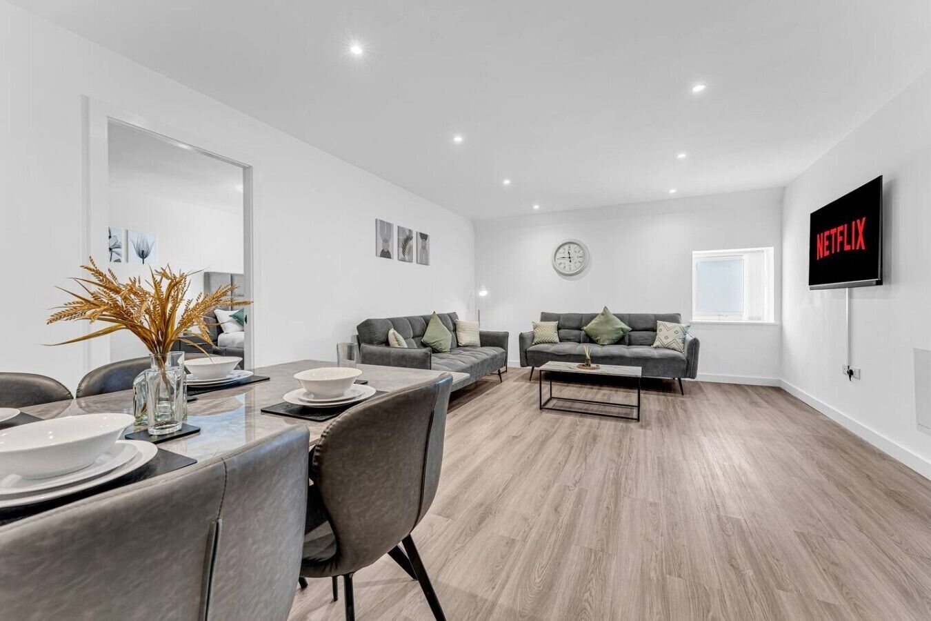 A modern living room with a dining area, a grey sofa set, a coffee table, and a flat-screen TV mounted on the wall.