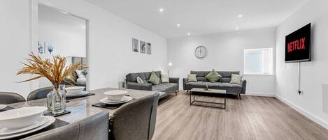 A modern living room with a dining area, a grey sofa set, a coffee table, and a flat-screen TV mounted on the wall.