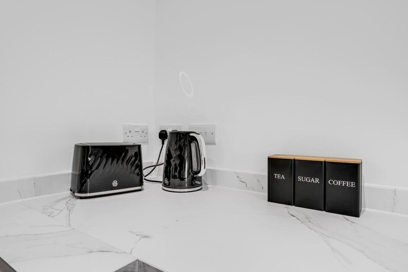 A kitchen counter with a toaster, a kettle, and three black boxes labeled tea, sugar, and coffee.