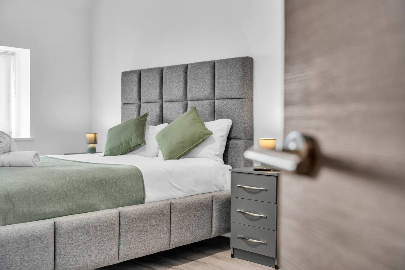 A modern bedroom with a grey upholstered bed, green pillows, a bedside table with a lamp, and a grey nightstand.