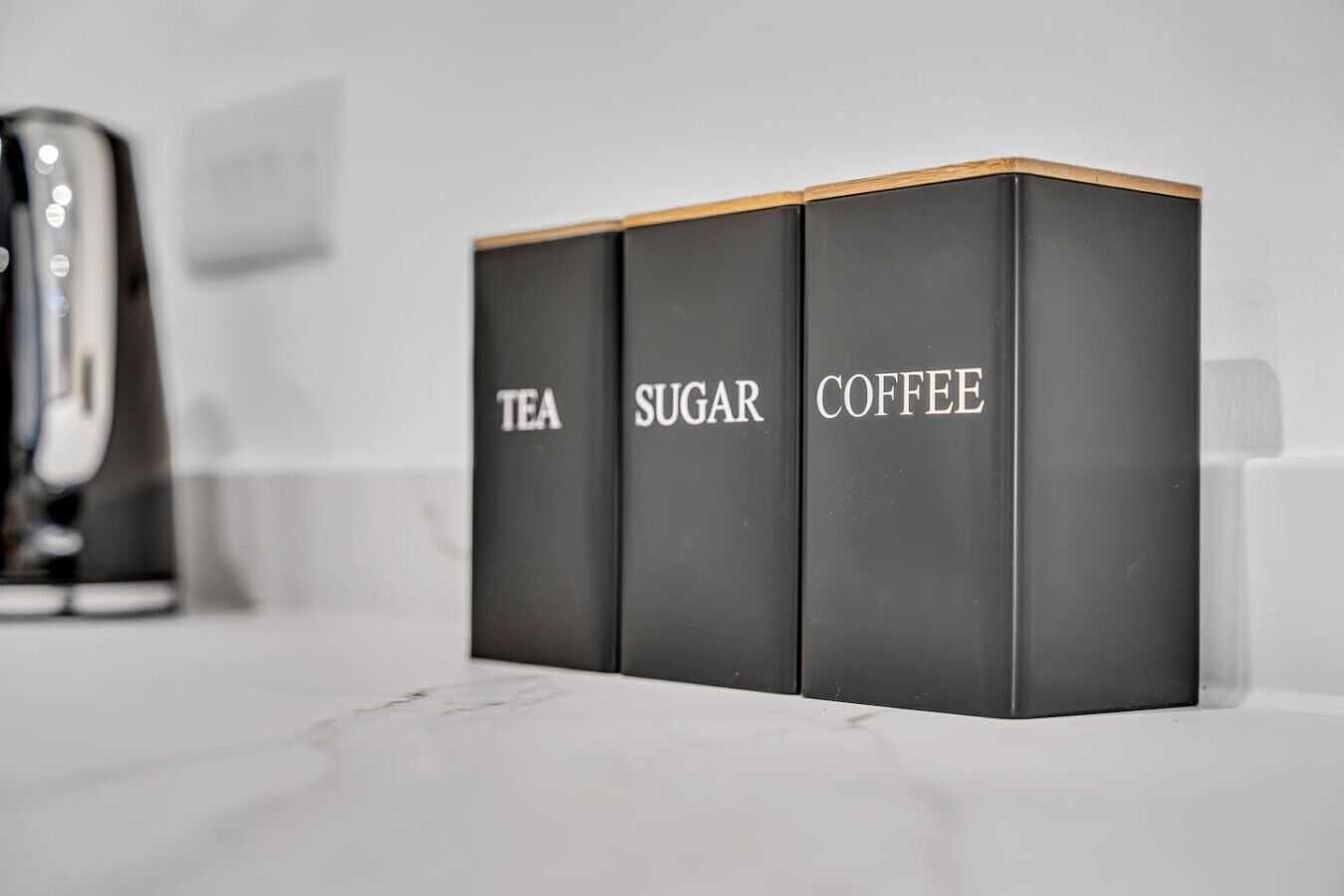 Un piano di lavoro in cucina con tre scatole nere contenenti tè, zucchero e caffè.
