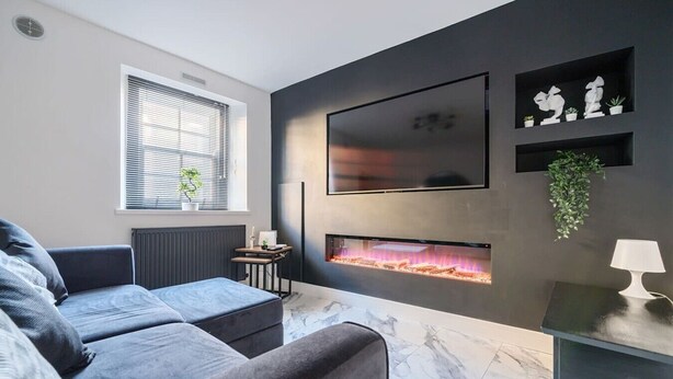 En moderne stue med en grå sofa, et fladskærms-tv monteret på væggen, en pejs og et lille bord med en lampe.