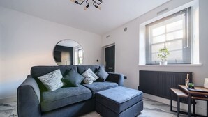 En moderne stue med en grå hjørnesofa, et rundt spejl, et spisebord med stole og et vindue med persienner.