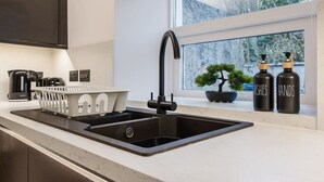 Et moderne køkken med en sort vask, en elkedel, et opvaskestativ og to flasker mærket 'DISHES' og 'HANDS' på vindueskarmen.