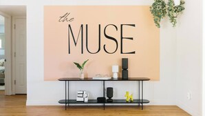 Huoneessa on suuri seinälle kiinnitetty kyltti, jossa lukee »THE MUSE», musta konsolipöytä koriste-esineineen ja roikkuva kasvi.
