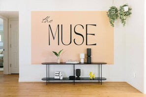 Ein Zimmer mit einem großen, an der Wand befestigten Schild mit dem Schriftzug 'the MUSE', einem schwarzen Konsolentisch mit Dekoration und einer hängenden Pflanze.